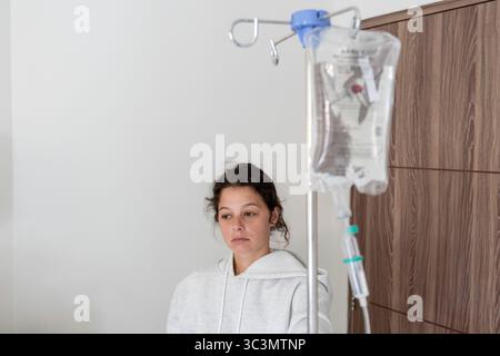 Une jeune femme a l'air malade, allongée dans un lit d'hôpital à Bali. Une perfusion intraveineuse se trouve à proximité, indiquant un traitement médical. La chambre est lumineuse et minimalement déco Banque D'Images