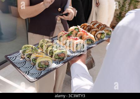 Serveur taillé méconnaissable tenant un plateau avec divers rouleaux de sushi, avec des ingrédients comme l'avocat, les asperges, le saumon, et nappé de sauce Banque D'Images