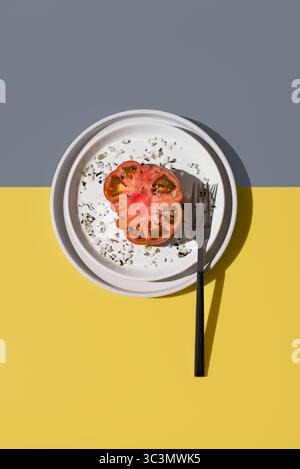 Vue de dessus d'une tomate raf coupée en deux, saupoudrée de sel et d'épices, élégamment placée sur une assiette blanche avec fourchette. Le backgrou jaune vibrant et gay Banque D'Images