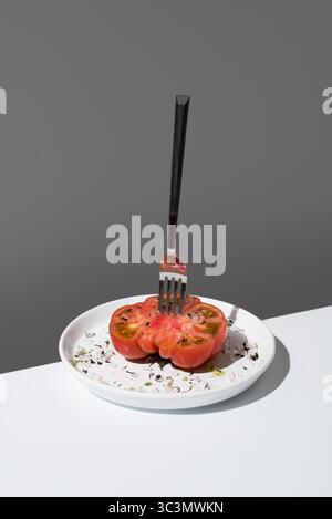 La moitié juteuse d'une tomate Raf assaisonnée de sel et d'épices repose élégamment sur une assiette blanche, avec fourchette. Les couleurs éclatantes contrastent avec le fond gris Banque D'Images
