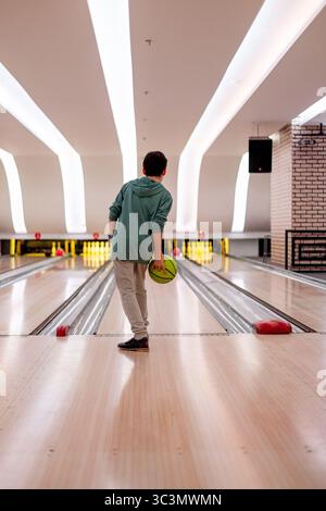 Un garçon se concentre intensément alors qu'il se prépare à jouer dans une piste de bowling moderne et élégante avec des plafonniers brillants et des couloirs laminés qui s'étendent jusqu'à l'intérieur Banque D'Images