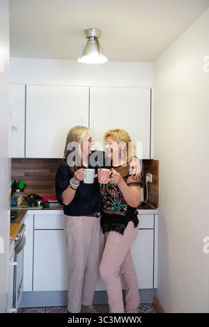 Deux amies seniors partagent un moment joyeux dans une cuisine confortable, riant et tenant des tasses. Leurs expressions joyeuses reflètent la chaleur, l'amitié, un Banque D'Images