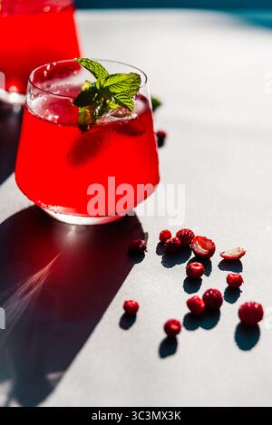 Verre rafraîchissant de fraises et de thé hibiscus est assis à la lumière directe du soleil, garni de menthe, entouré de baies fraîches dispersées sur un surf lumineux Banque D'Images