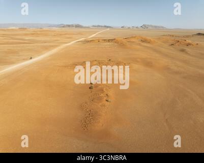 Vue aérienne d'une route sablonneuse traversant l'étendue dorée du désert du Namib vers des montagnes lointaines, Walvis Bay, Walvis Bay, Namibie. Banque D'Images