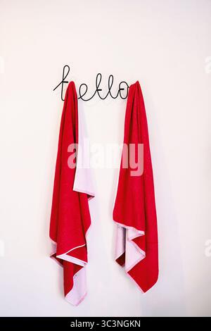 Deux serviettes de plage rouges suspendues à un cintre dans un hôtel. Le cintre est sous la forme de l'inscription Hello. Serviettes contre un mur blanc. Banque D'Images