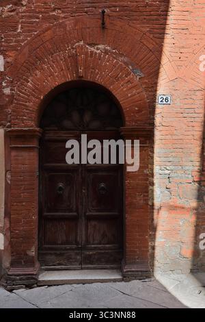 134 porte en arc rond découpée dans la façade en briques rouges d'une maison de ville sur la rue via Mazzini où le soleil projette des jeux de soleil et d'ombre. Montalcino-Toscane-Italie Banque D'Images
