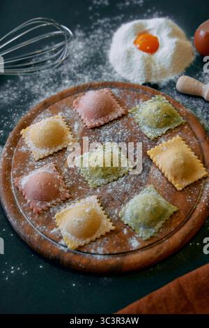 Raviolis faits maison colorés sur une planche en bois avec de la farine et du jaune d'œuf entouré d'outils de cuisine et d'ingrédients pour une expérience culinaire délicieuse Banque D'Images