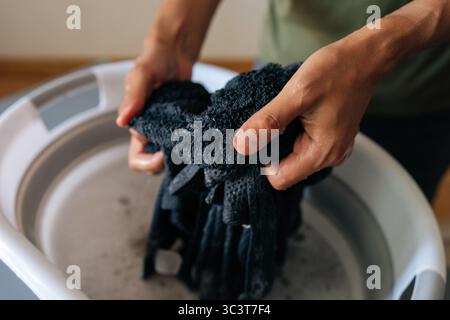 Plan recadré d'une personne méconnaissable trempant du linge noir dans l'eau dans un bassin pliant debout à une table grise faisant des tâches ménagères, gros plan. Banque D'Images