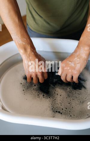 Plan vertical recadré d'un homme méconnaissable trempant du linge noir dans l'eau dans un bassin pliant debout à une table grise faisant des tâches ménagères. Banque D'Images