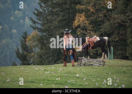 Une femme en train occidental s'approche d'un cheval noir sellé sur une colline herbeuse près des arbres. Elle porte un chapeau de cow-boy, des chaps et des bottes à talons. Banque D'Images