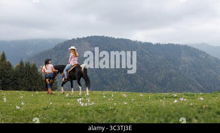 Une femme blonde monte un cheval noir et blanc sur un terrain herbeux avec un fond de montagne. Une autre femme dirige. Banque D'Images