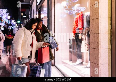 Deux jeunes femmes regardent à travers une vitrine festive de magasin présentant des mannequins dans des tenues saisonnières tout en tenant des sacs à provisions pendant une soirée cit Banque D'Images