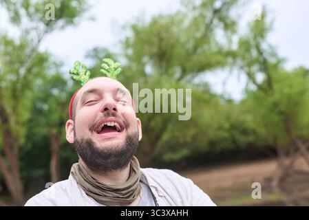 Un jeune homme hispanique portant un serre-tête drôle de renne de Noël en bois sourit avec une expression drôle à l'extérieur, dans un cadre naturel. Banque D'Images