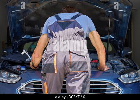 Vue arrière du mécanicien masculin debout près du capot de voiture ouvert avec la clé à la main. Homme en salopettes de travail réparant le moteur de voiture dans le garage Banque D'Images