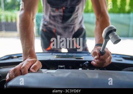 Mécanicien masculin utilisant une clé à douille pour réparer le moteur de voiture dans le garage. Homme avec des outils de réparation dans les mains sous le capot ouvert Banque D'Images
