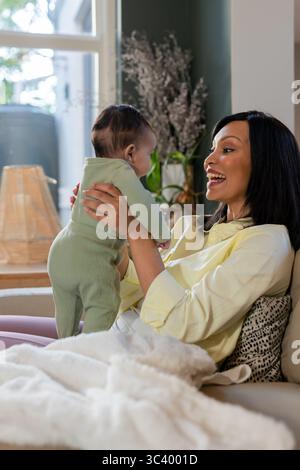 Mère et fille tenant bébé debout sur le canapé à la maison, souriant à côté de la lampe en osier, couverture crème Banque D'Images