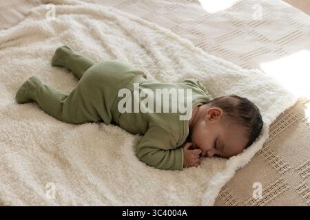 Bébé fille portant une combinaison verte couchée face vers le bas sur une couverture blanche sur jeté beige dans la chambre Banque D'Images