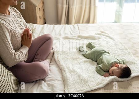 Mère médite à côté de bébé endormi sur une couverture blanche moelleuse à travers le lit de couette près de la fenêtre Banque D'Images