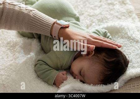 Mère portant un pull en tricot et smartwatch touchant les cheveux de la fille de bébé à la maison sur une couverture en peluche Banque D'Images