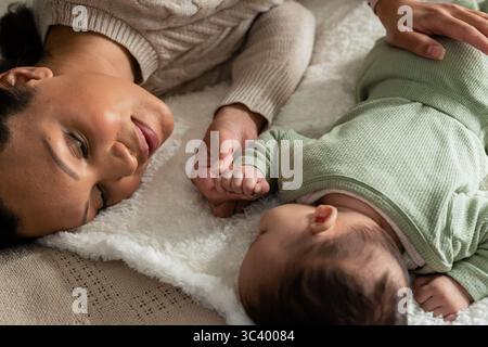 Mère en chandail en tricot câblé tenant bébé fille en combinaison couchée sur le lit sous la couverture de fausse fourrure Banque D'Images