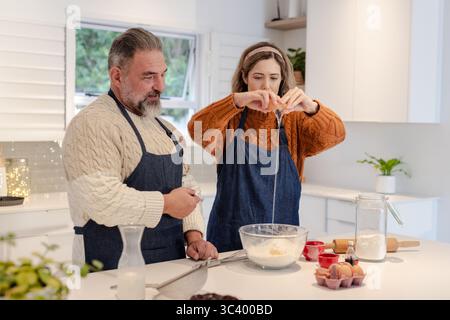 Couple portant des tabliers bleus cuisant à l'îlot de cuisine craquant l'œuf dans le bol tout en tenant le fouet Banque D'Images