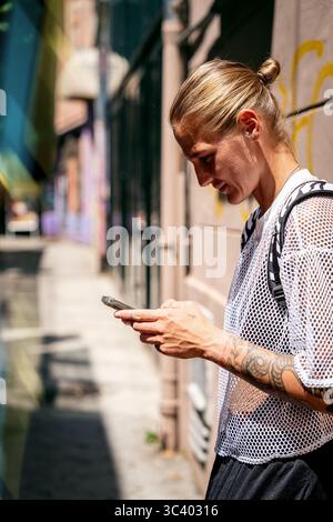Jeune femme urbaine dans le style de la rue vérifiant son smartphone dans la rue de la ville en été Banque D'Images