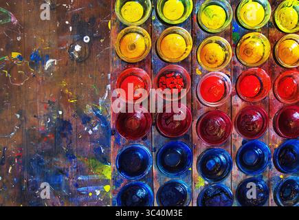 Une vue aérienne de bols de pigment en verre soigneusement disposés dans un spectre de couleurs complet jaune, rouge et bleu contraste avec un travail en bois salissant et éclaboussé Banque D'Images