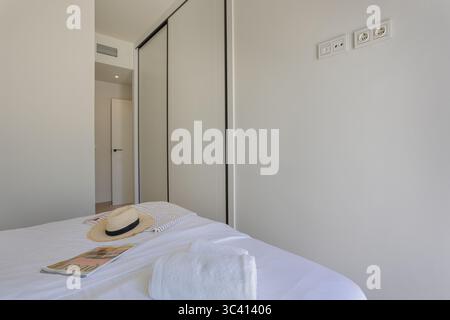 Chambre minimaliste avec draps blancs, chapeau de paille et serviette sur le lit, grande armoire intégrée et accessoires modernes. Banque D'Images