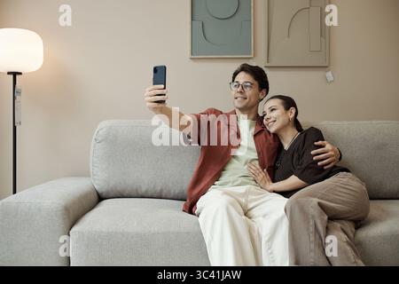 Jeune homme caucasien adulte assis sur le canapé prenant selfie avec smartphone, tandis que jeune femme caucasienne adulte assise à côté de lui embrassant et souriant dans la salle d'exposition du magasin de meubles Banque D'Images