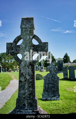 Une croix celtique altérée se dresse dans un cimetière irlandais, entourée d'anciennes pierres tombales sous un ciel bleu clair. Banque D'Images