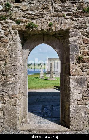 Vue à travers une arche d'un site historique irlandais, mettant en valeur un paysage tranquille avec une croix et des ruines d'église. Banque D'Images