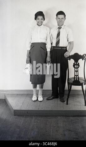 Années 1950, historiques, debout sur une plate-forme en bois dans un studio photographique... une jeune femme des caraïbes, sac à main dans sa main droite, main gauche tenant la main avec un homme anglais plus âgé, dans une chemise à manches courtes et cravate cachée dans son pantalon, alors qu'ils posent ensemble pour une photo de portrait, Angleterre, Royaume-Uni. Banque D'Images