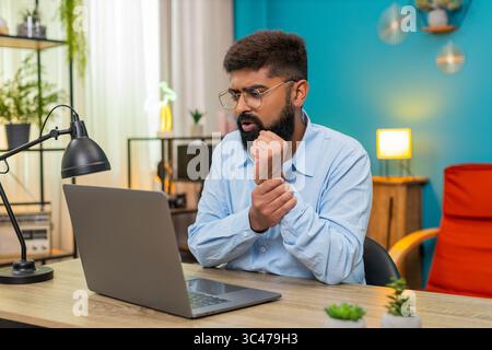 Homme indien à la maison massages poignet douloureux après une longue session d'ordinateur portable mal de tension répétitive. Le gars arabe au bureau fléchit les doigts sent la douleur chercher le soulagement avec des exercices doux en espérant que les tendons récupèrent. Banque D'Images