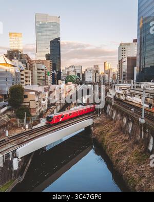 Chiyoda City, Japon - 03 juin 2025 : vue aérienne d'un train rouge éclatant traversant le paysage urbain près du pont Hijiri, reflété dans la rivière calme en contrebas. Banque D'Images