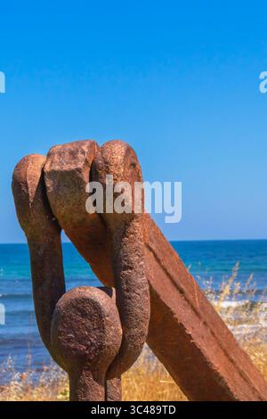 Une grande chaîne en métal rouillé, peut-être une chaîne d'amarrage, repose sur une zone herbeuse près du rivage. La mer Noire scintille en arrière-plan sous un nuage Banque D'Images