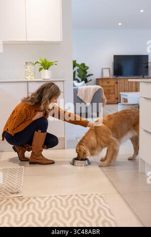 Femme agenouillée sur le sol de la cuisine tapotant Golden retriever mangeant au bol en acier, copiez l'espace Banque D'Images
