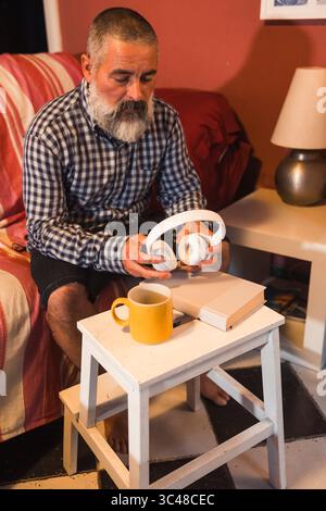 Homme senior détendu tenant des écouteurs, dégustant une tasse de thé ou de café, avec un livre sur la table d'appoint dans son salon Banque D'Images