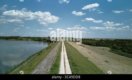 Vue aérienne panoramique d'une route rurale de gravier longeant le bord du lac Pavlovac en Serbie. Entouré de terres agricoles et de douces collines sous un clair Banque D'Images