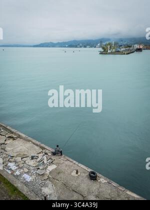 Vue aérienne d'un pêcheur solitaire jetant sa ligne de la jetée en béton altéré dans les eaux tranquilles et turquoises près du port lointain, Batoumi, Adjara, Géorgie. Banque D'Images