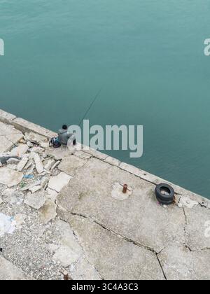 Vue aérienne d'un pêcheur solitaire jetant sa ligne dans la mer tranquille et Azur depuis une jetée en béton altérée, Batoumi, Adjara, Géorgie. Banque D'Images