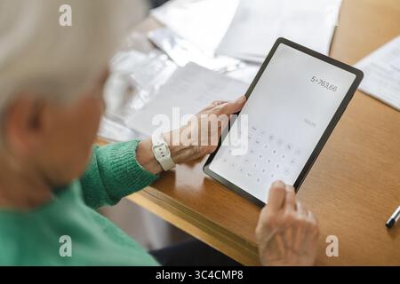 Femme senior utilisant l'application calculatrice sur tablette numérique à table Banque D'Images