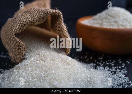 Sucre blanc renversé d'un sac de toile de jute à côté d'un bol plein, placé sur un fond sombre. Parfait pour l'emballage alimentaire, les visuels culinaires ou la traditiona Banque D'Images