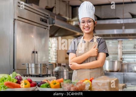 Jeune femme chef souriante et montrant un geste de pouce levé dans un environnement de cuisine professionnel. Elle porte un tablier rayé et un chapeau de chef blanc, Conve Banque D'Images