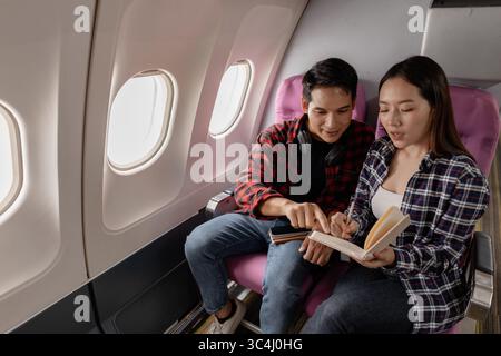 Jeune couple asiatique assis ensemble sur des sièges d'avion, discutant de plans de voyage et écrivant des notes dans un cahier pendant leur vol. Ils sourient et enj Banque D'Images