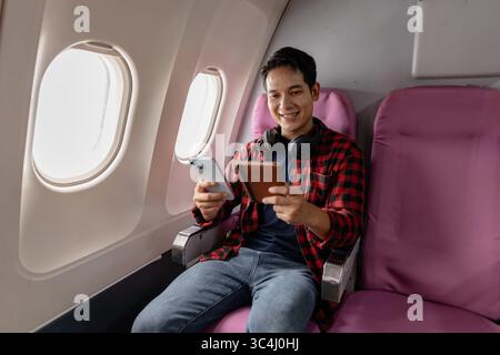 Un jeune homme est assis dans un avion, profitant de son vol tout en vérifiant son smartphone et son passeport. Il semble détendu et content, reflétant le moderne Banque D'Images