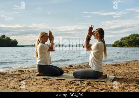 Deux personnes sont engagées dans une séance de yoga sur une plage de sable au bord d'un lac tranquille. Le soleil se couche, projetant des couleurs douces à travers le ciel. Ils exécutent un moi Banque D'Images