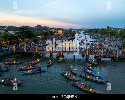 Vue aérienne des bateaux ornés de lanternes flottent sereinement sur la rivière, reflétant la lueur chaude du coucher du soleil et le pont animé, Hội an, Quảng Nam, Vietnam. Banque D'Images