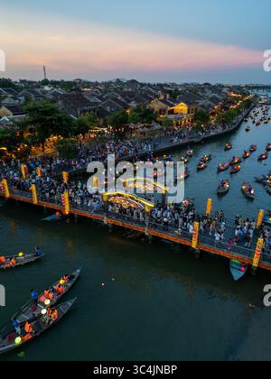 Vue aérienne d'un pont éclairé par des lumières chaudes, animé par des gens et des bateaux dérivant le long de la rivière, reflétant le ciel crépusculaire, Hội an, Quảng Nam, Vietnam. Banque D'Images
