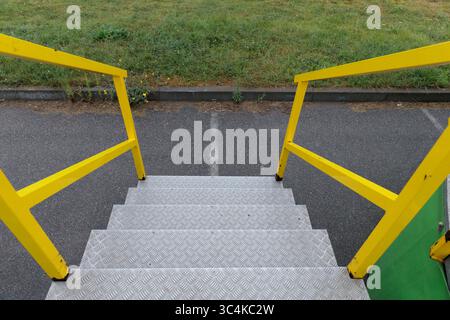 Vue rapprochée de balustrades jaunes et de marches métalliques menant d'une structure à un sentier asphalté avec de l'herbe et des fleurs sauvages en arrière-plan Banque D'Images