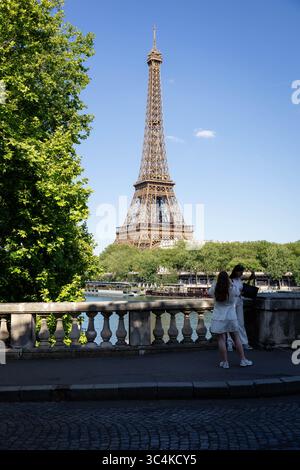 Vue en bas angle sur la Tour Eiffel, deux femmes en blanc prenant une photo, arbres verdoyants et balustrade en pierre. Ambiance lumineuse et ensoleillée ; idéal pour les voyages Banque D'Images